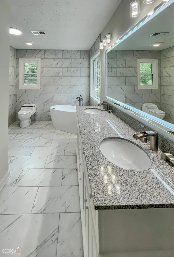 Completed bathroom remodel with a double-sink vanity, granite countertop, large mirror, freestanding soaking tub, and white marble-look tile covering the floor and walls.