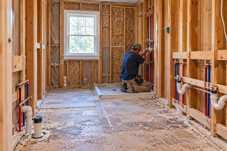 A contractor works on exposed plumbing in a bathroom that has been gutted down to the studs.
